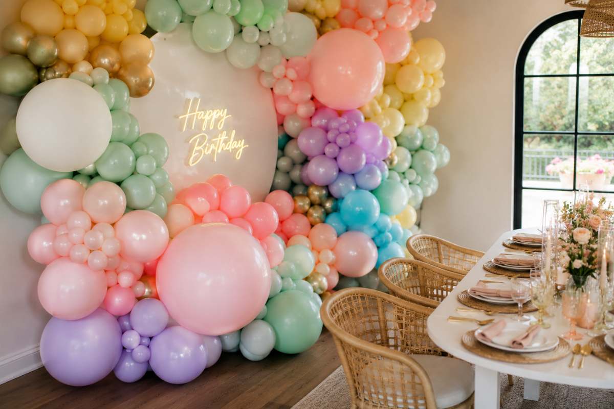 Colorful balloon installation featuring oversized pastel and metallic balloon clusters arranged around a modern birthday backdrop with a neon sign, creating a stunning photo-worthy focal point beside an elegantly styled party table.
