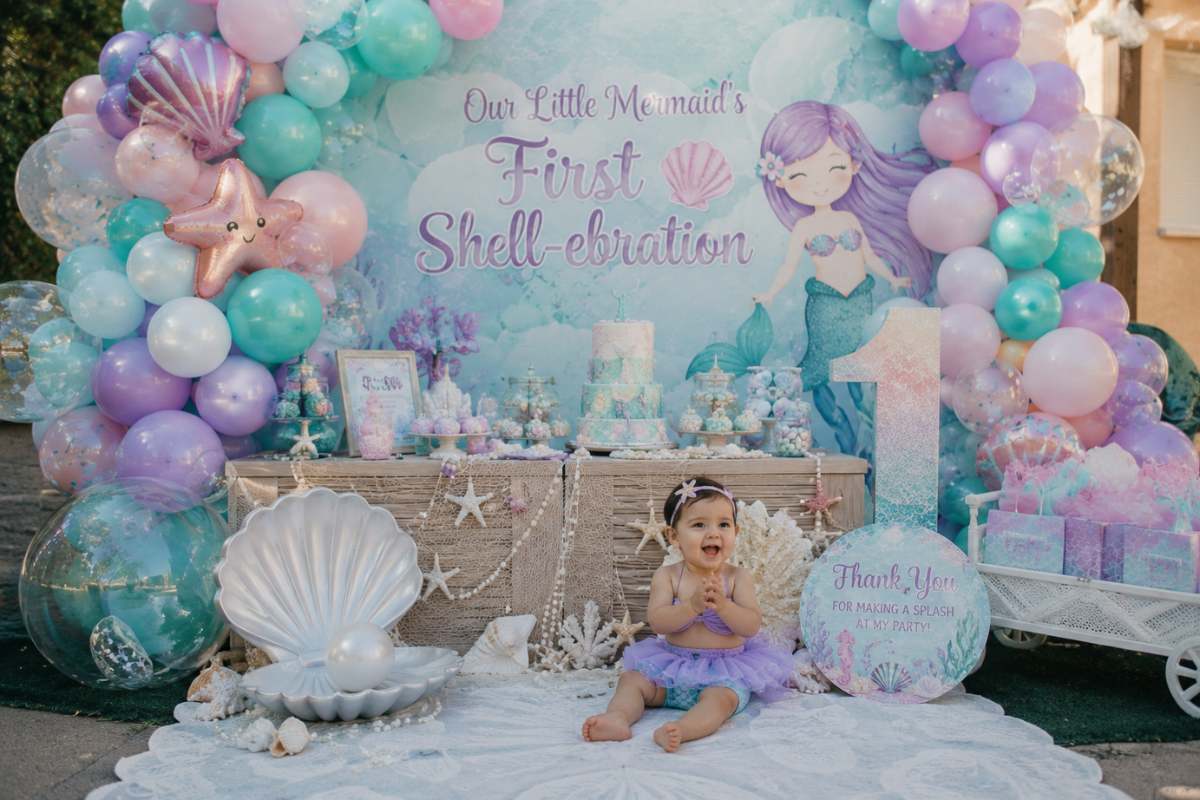Mermaid-themed first birthday party featuring a pastel teal, lavender, and pink balloon backdrop, seashell decorations, pearl accents, ocean-inspired dessert table, mermaid cake, starfish cookies, oversized shell prop, and a baby girl celebrating her First Shell-ebration in a magical underwater-inspired setting.