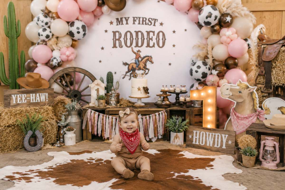 Vertical My First Rodeo first birthday party setup featuring a western-themed backdrop with pastel pink and brown balloon garland, cow-print accents, hay bales, cowboy hat decorations, rustic dessert table, cactus props, light-up number one sign, cowhide rug, and a smiling baby dressed in a cowboy-inspired outfit.