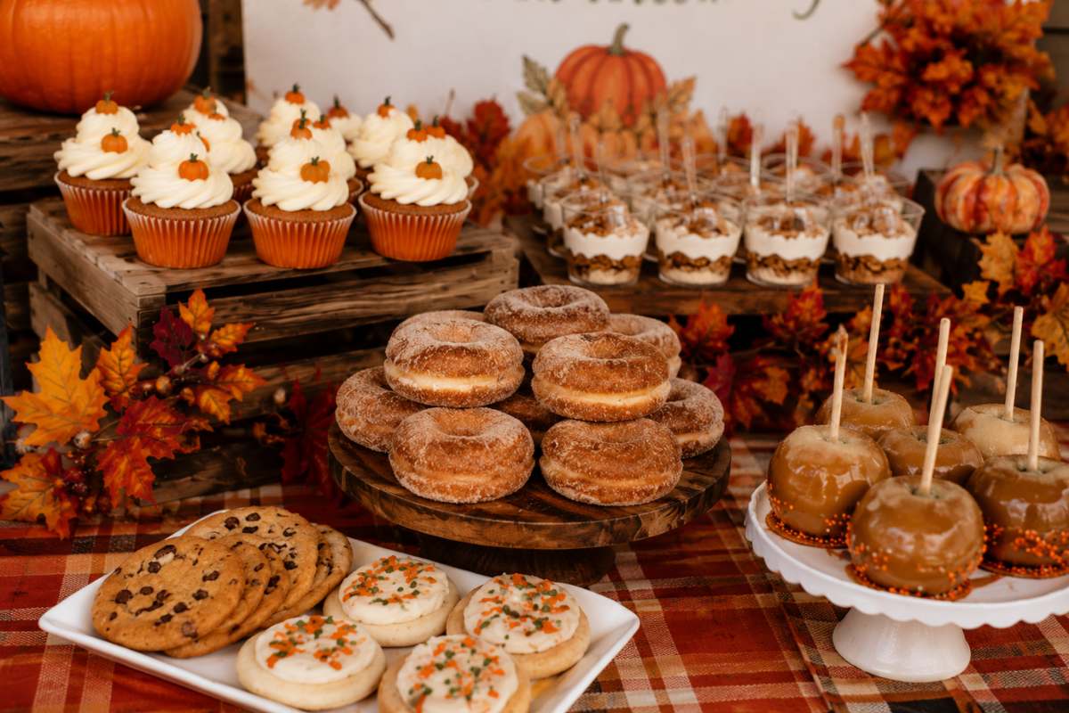 Close-up of a pumpkin patch baby shower dessert table featuring pumpkin spice cupcakes, apple cider donuts, caramel apples, frosted fall cookies, parfaits, and rustic autumn decorations with pumpkins, plaid linens, and colorful fall leaves.