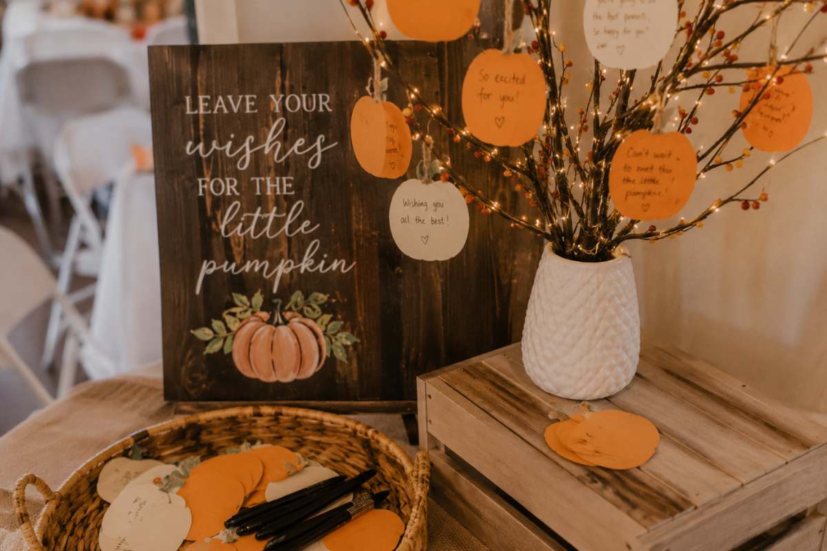 Pumpkin patch baby shower guestbook alternative featuring a rustic wish tree decorated with handwritten pumpkin-shaped message cards, a wooden sign inviting guests to leave wishes for the little pumpkin, and a basket of blank cards and pens for guests to share heartfelt notes.