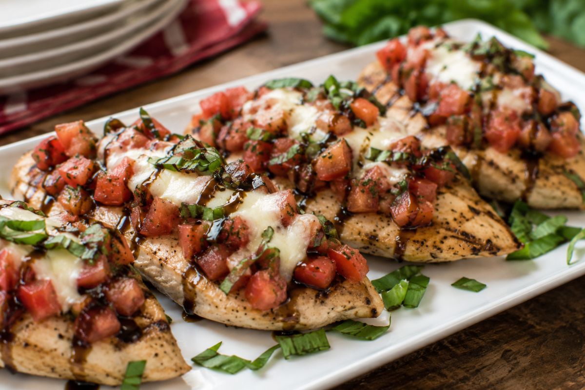 Grilled bruschetta-inspired chicken topped with diced tomatoes, fresh basil, melted mozzarella, and balsamic glaze, served on a white platter as an elegant and budget-friendly wedding reception entrée.