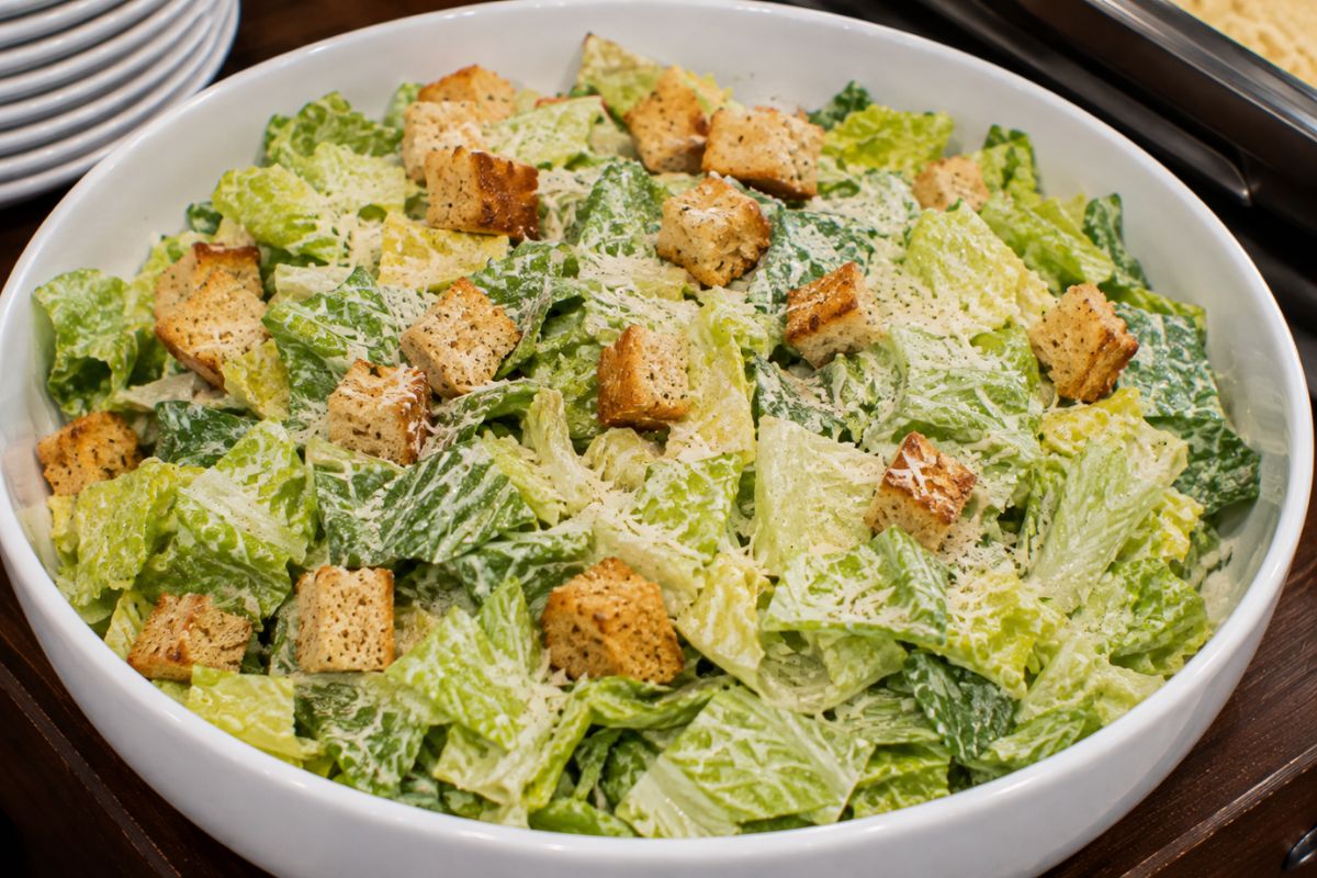 Classic Caesar salad with crisp romaine lettuce, crunchy croutons, freshly grated Parmesan, and creamy Caesar dressing served in a large white bowl as a fresh and budget-friendly side dish for a wedding reception buffet.