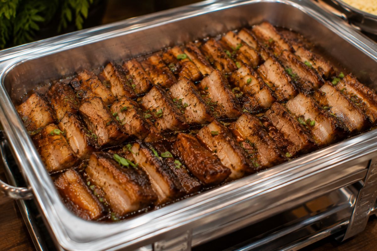 Glazed pork belly sliced into bite-sized portions with caramelized edges and a honey garlic herb glaze, served in a stainless steel buffet chafing dish as an elegant and budget-friendly wedding reception entrée.