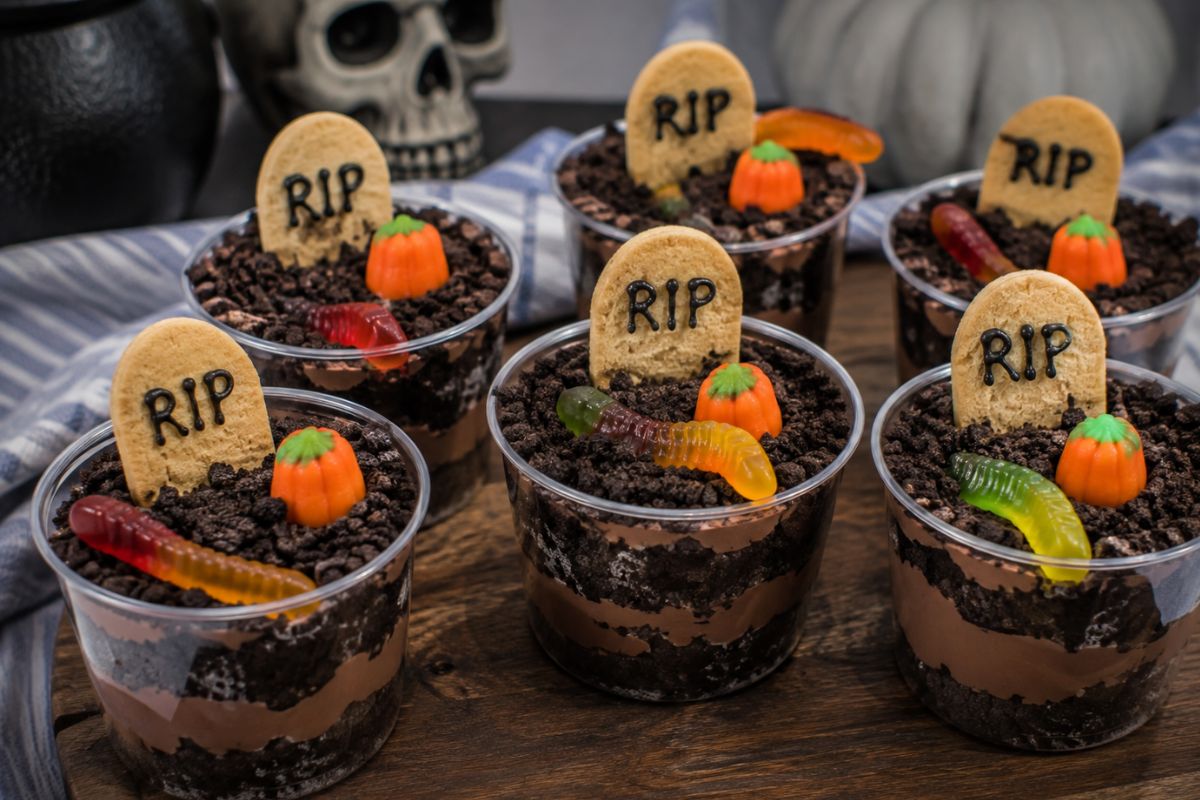 Graveyard dessert cups layered with chocolate pudding and crushed chocolate cookies, topped with cookie tombstones, candy pumpkins, and gummy worms.