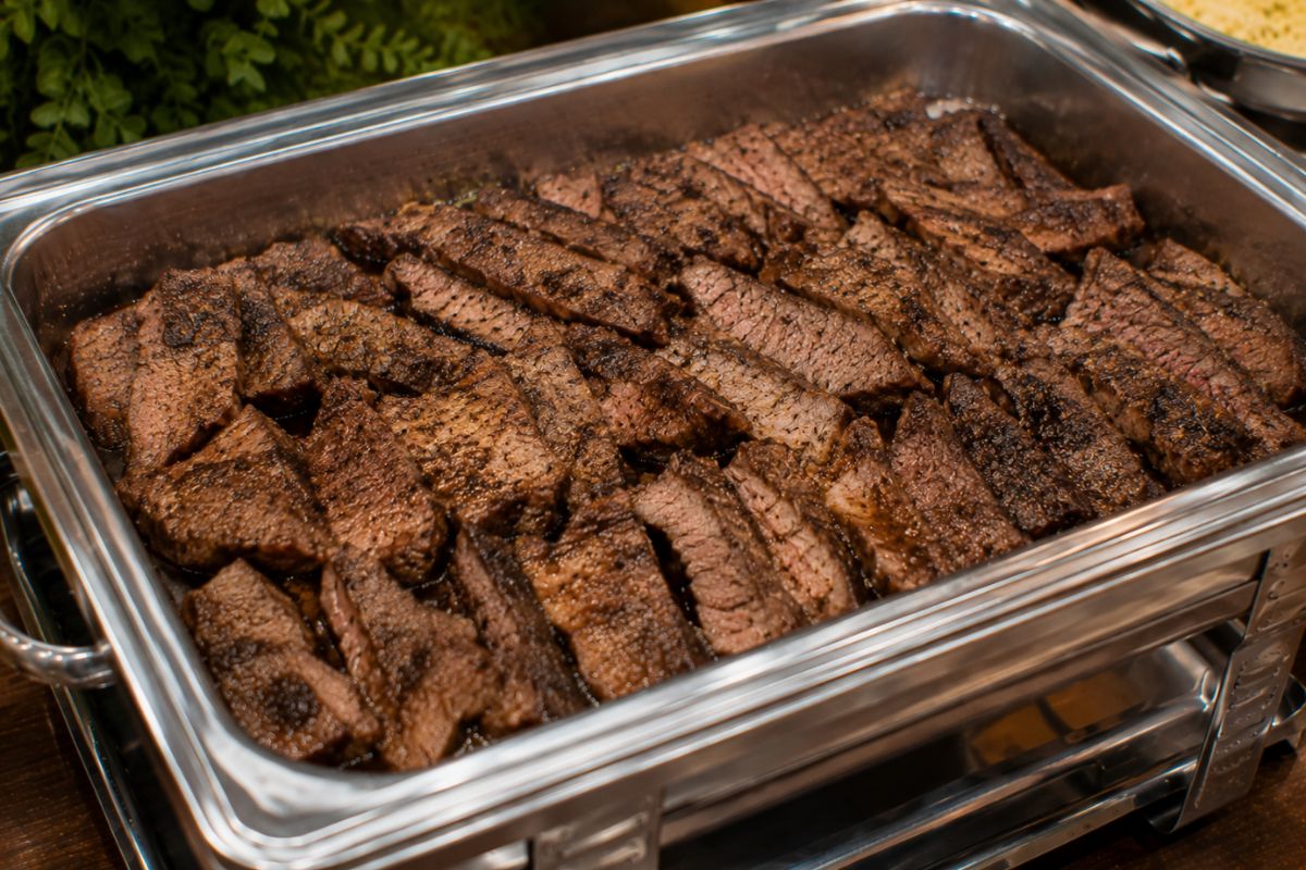 Thinly sliced grilled seasoned steak with a flavorful herb and garlic crust, arranged in a stainless steel buffet chafing dish as an elegant and budget-friendly wedding reception entrée.