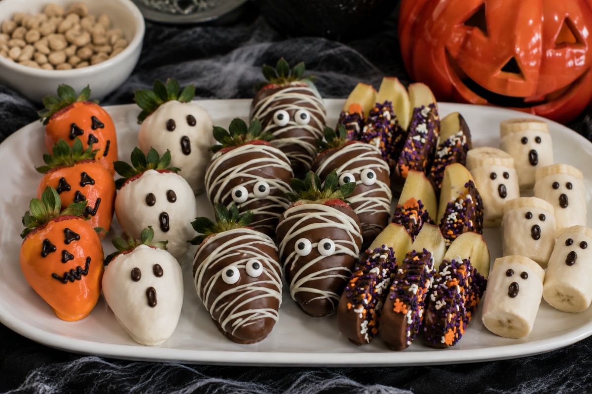Halloween chocolate-dipped fruit treats featuring decorated strawberries, chocolate-covered apple slices, bananas, candy eyes, colorful drizzle, and festive sprinkles.