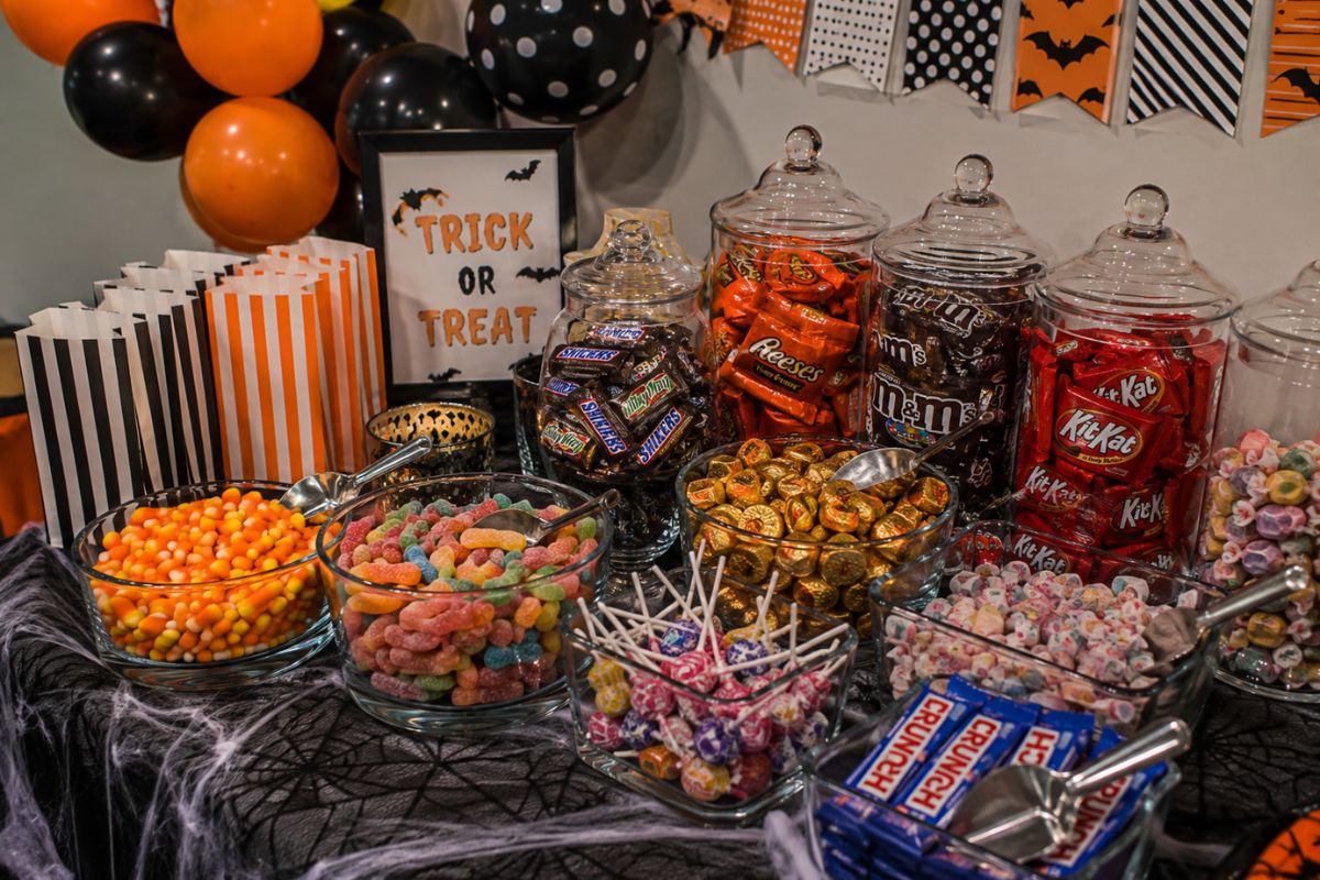 Halloween self-serve candy station with glass jars, bowls, scoops, chocolate bars, gummy candies, candy corn, lollipops, and wrapped sweets.