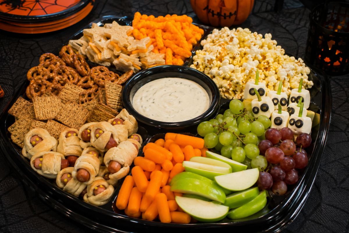 Halloween snack tray with pretzels, crackers, popcorn, chips, mummy sausage bites, fresh fruit, vegetables, dip, and bite-sized treats arranged for easy party snacking.