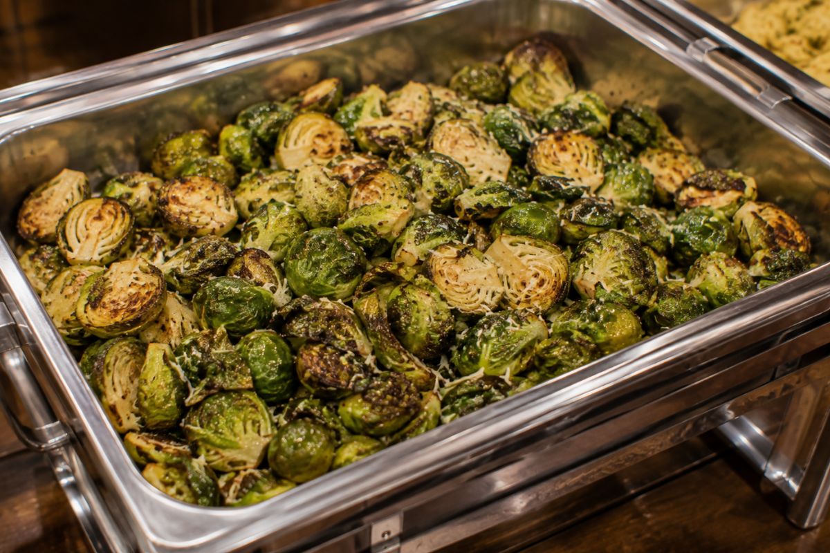 Oven-roasted Brussels sprouts with crispy caramelized edges, lightly seasoned with herbs and grated Parmesan, served in a stainless steel buffet chafing dish as an elegant and budget-friendly wedding reception side dish.