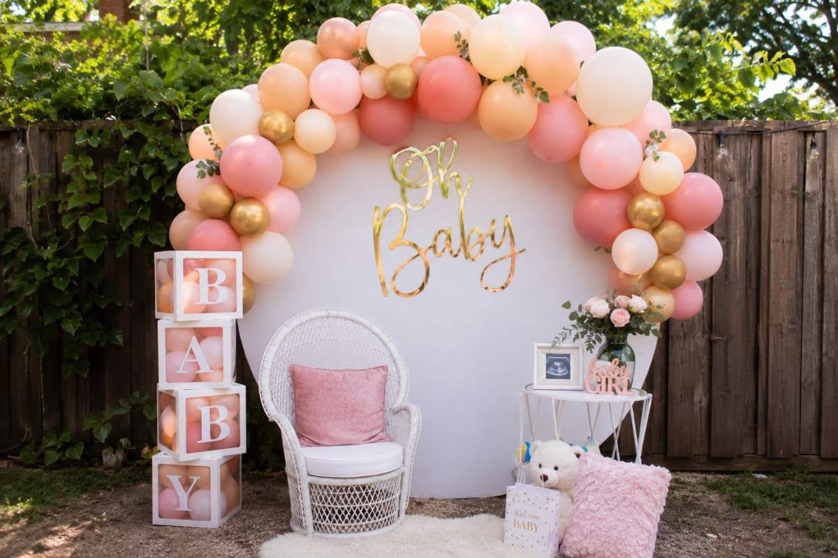 Elegant DIY balloon garland backdrop with blush, peach, white, and gold balloons, an "Oh Baby" sign, wicker chair, BABY blocks, and floral accents in a backyard baby shower setting.