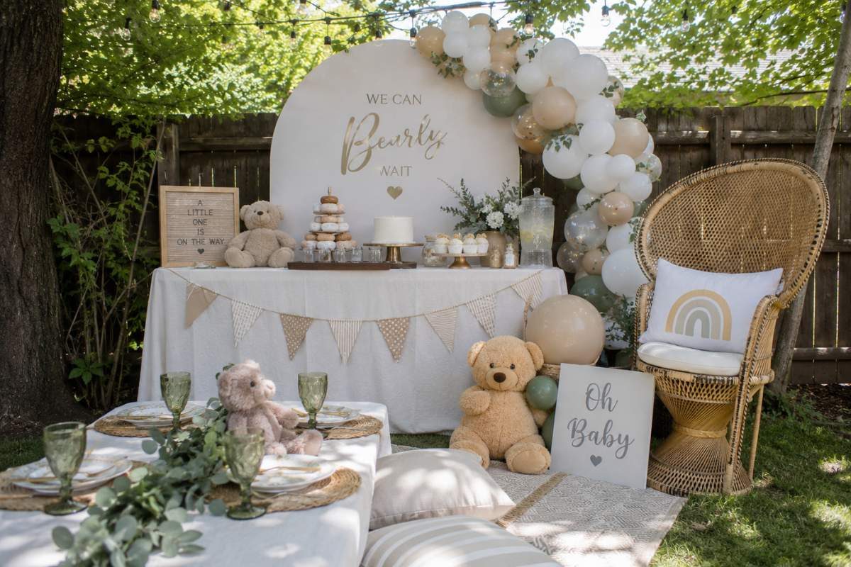 Gender-neutral backyard baby shower featuring beige and white balloon garlands, teddy bear decorations, boho picnic seating, wicker chair, neutral dessert table, and soft earth-tone décor.