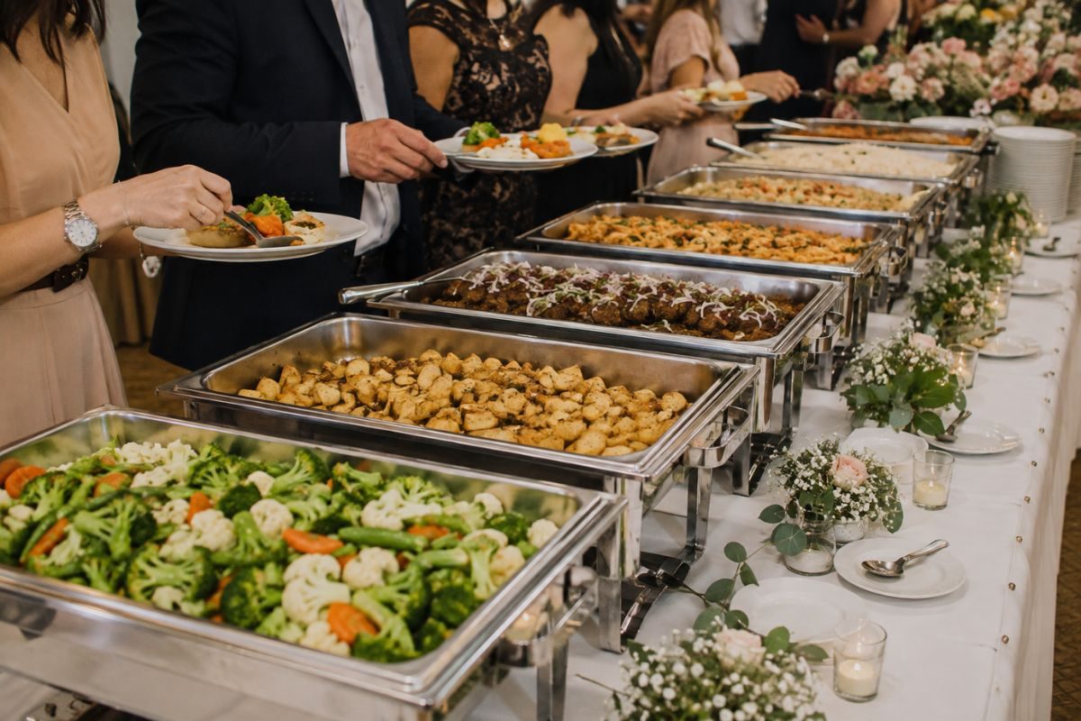 Wedding guests serving themselves from an elegant buffet featuring a variety of hot entrées, vegetables, pasta, and side dishes arranged on a beautifully decorated reception table with floral centerpieces and candles.