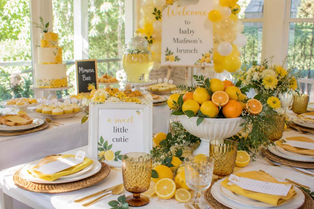 Citrus-themed brunch baby shower décor featuring lemons, oranges, yellow floral arrangements, balloon accents, citrus centerpieces, and elegant brunch table settings in a bright sunlit venue.