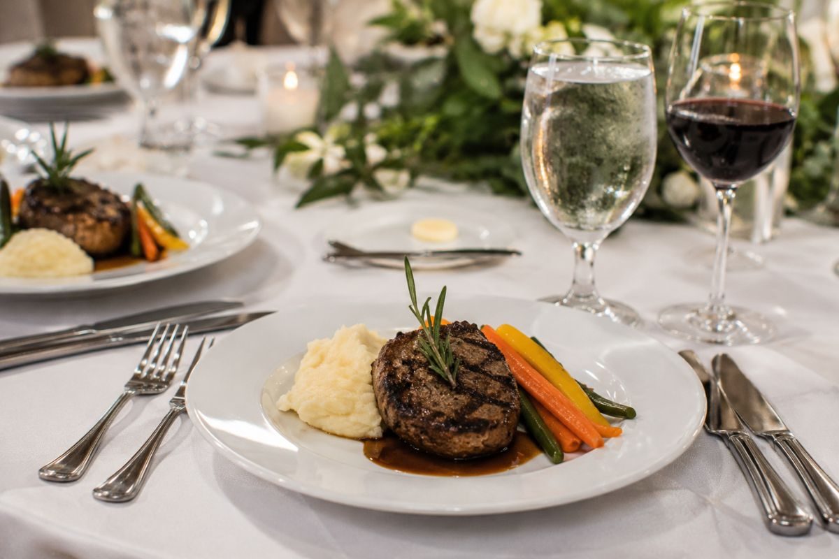 Elegant plated wedding dinner featuring grilled steak with creamy mashed potatoes, seasonal vegetables, and rosemary garnish on a white plate, set on a beautifully decorated reception table with glassware and floral centerpieces.