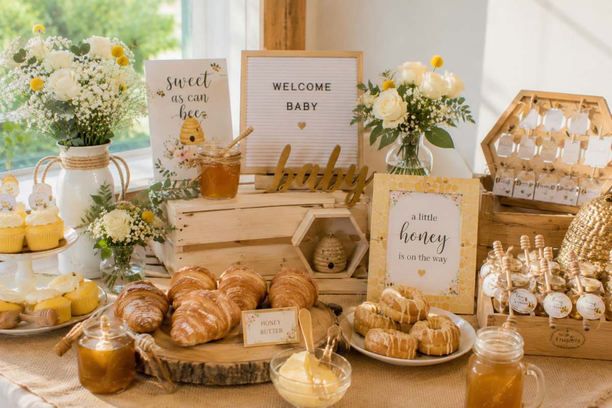 Honey-inspired brunch baby shower display featuring honey jars, croissants, glazed pastries, bee-themed decorations, fresh white flowers, honey butter, and warm golden décor in a cozy celebration setting.