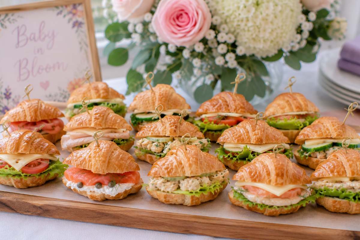 Mini croissant sandwiches filled with smoked salmon, turkey, chicken salad, and fresh vegetables arranged on a wooden serving board for a baby shower brunch.