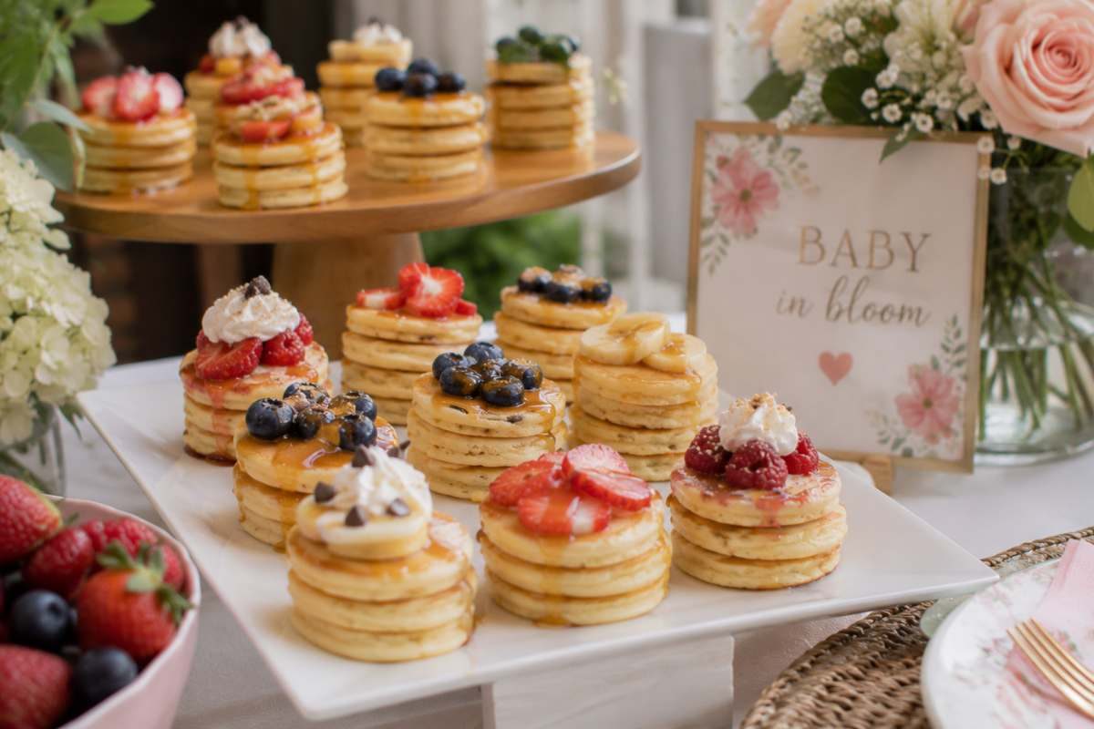 Mini pancake stacks displayed at a brunch baby shower, topped with fresh strawberries, blueberries, bananas, whipped cream, chocolate chips, and maple syrup alongside soft floral décor.