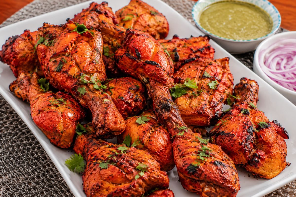 Indian-style tandoori chicken with charred edges and vibrant spices, garnished with fresh cilantro and served with mint chutney and sliced red onions, making a flavorful and budget-friendly wedding reception entrée.